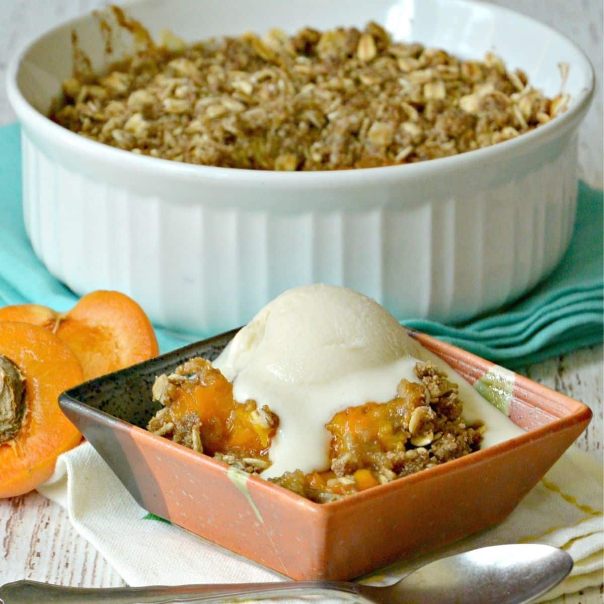 Small bowl with serving of apricot crisp topped with vanilla ice cream with fresh apricot and baking dish of dessert in the background