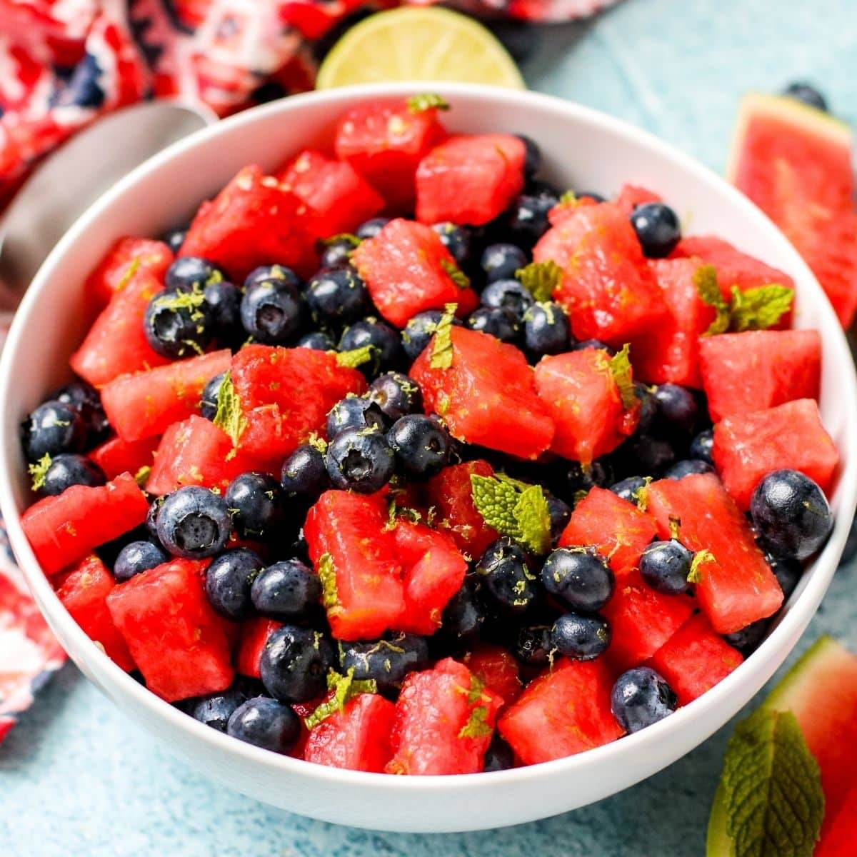 Bowl of fruit salad surrounded by slice of lime, watermelon wedges, and fresh mint leaves