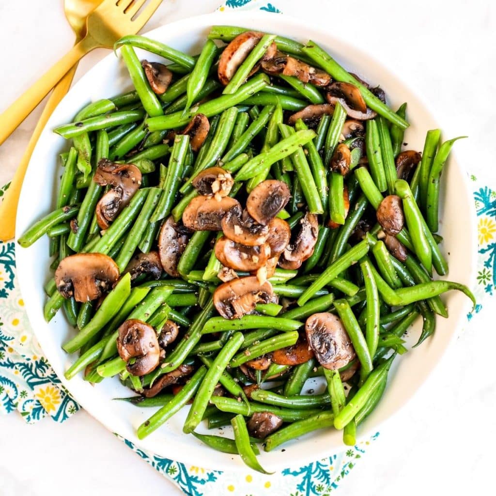 Serving bowl of green beans and mushrooms with gold utensils