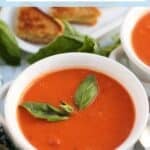 Bowl of tomato soup garnished with fresh basil with grilled cheese sandwiches in the background, with text overlay: Homemade Creamy Vegan Tomato Soup.
