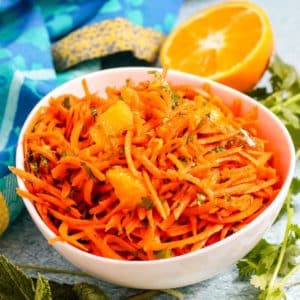 Bowl of carrot and orange salad surrounded by sprigs of fresh cilantro and mint