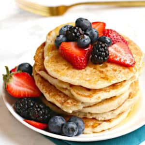 Stack of pancakes topped with fresh berries on a white plate with a gold fork in the background