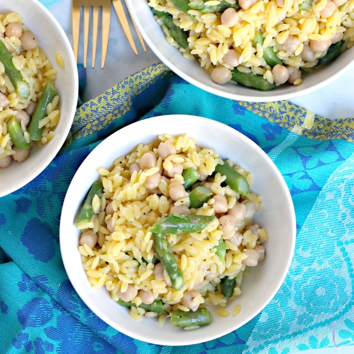 Overhead of bowls of lemon orzo with asparagus and chickpeas with a blue napkin
