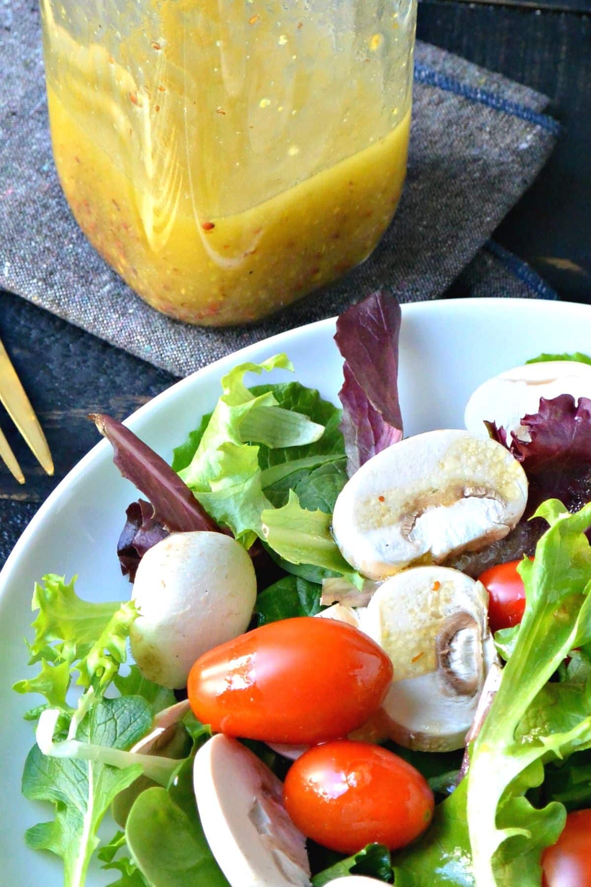 Plate of salad with mixed greens, mushrooms, and tomatoes with jar of Dijon vinaigrette behind it.