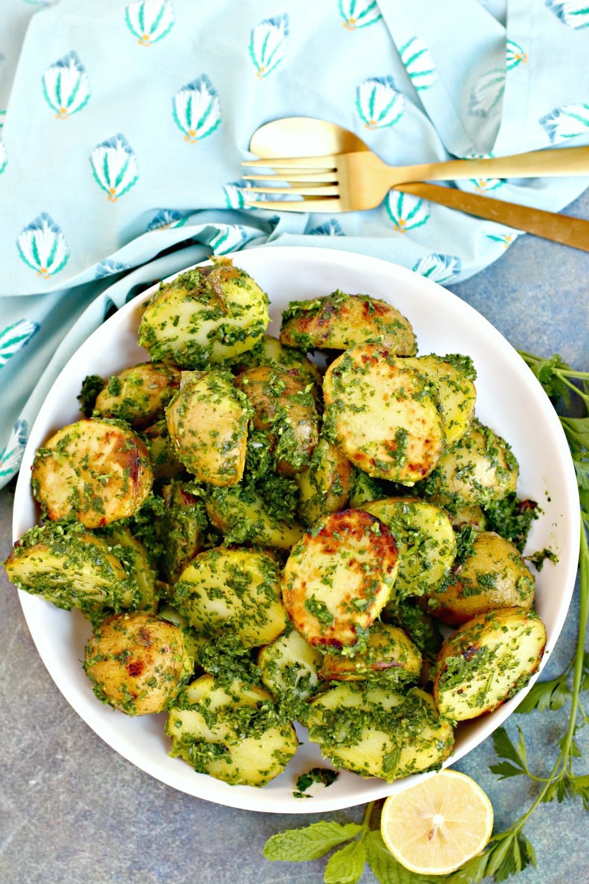 Grilled yellow potatoes in a bowl tossed with herb sauce next to aqua cloth napkin and gold serving utentils