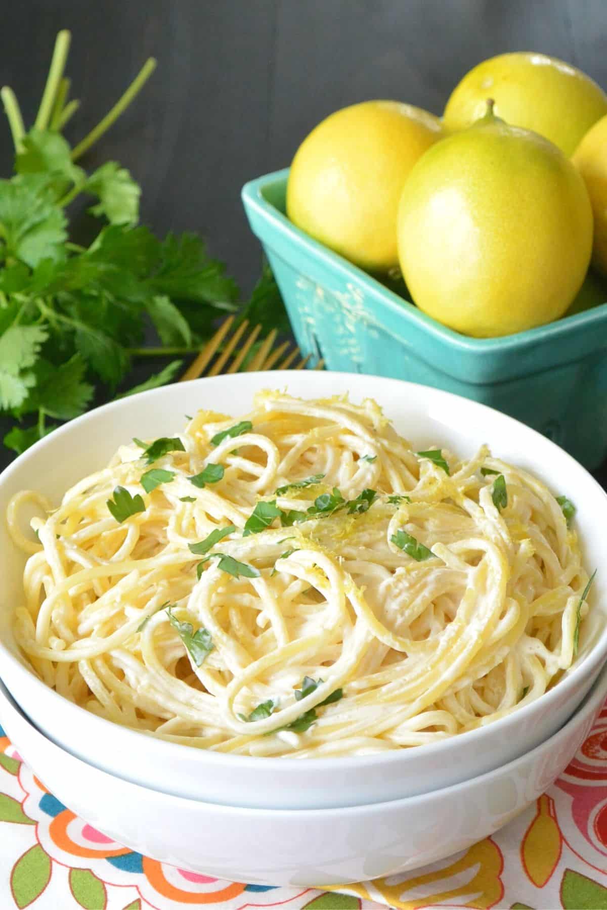 White bowl of pasta with aqua basket of lemons and bunch of parsley in the background