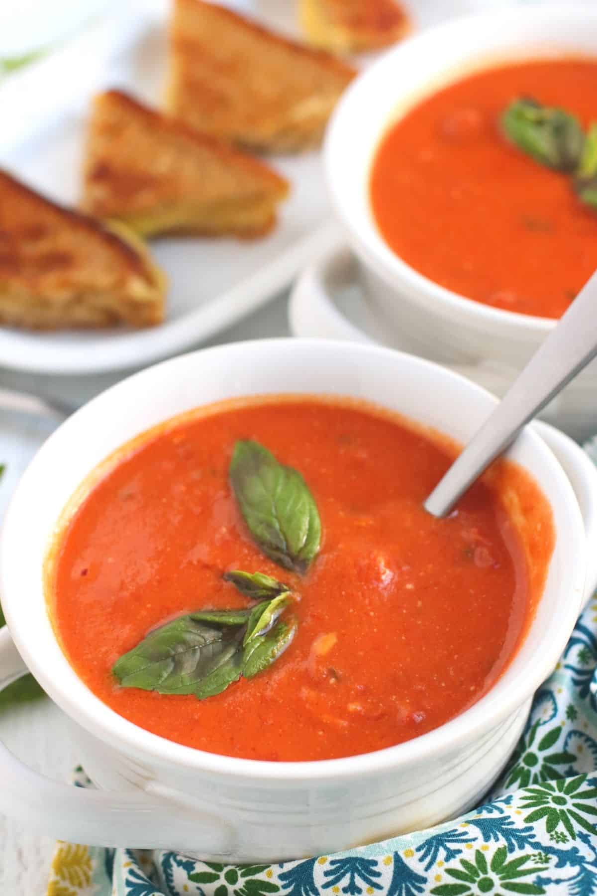 White bowls of creamy vegan tomato soup garnished with fresh basil with plate of grilled cheese sandwiches in the background.