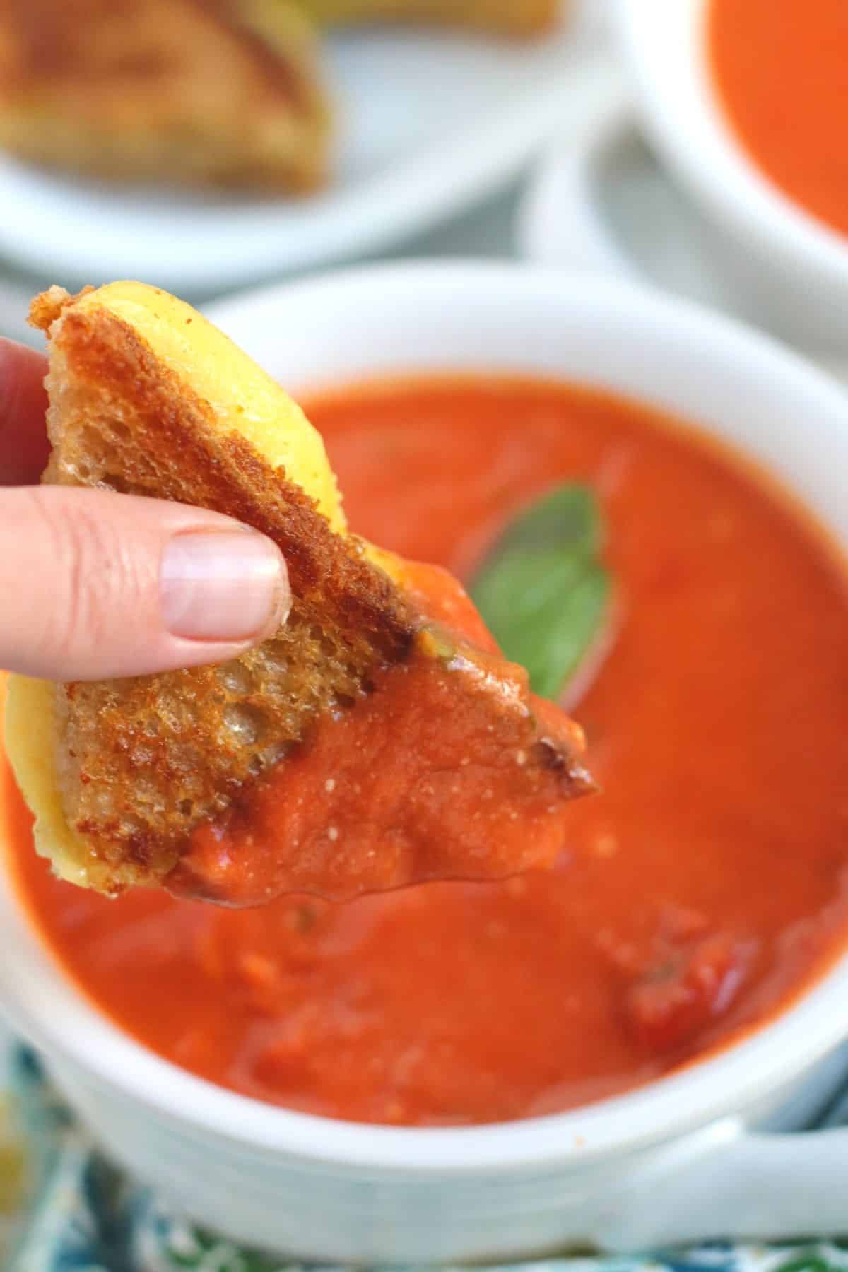 Hand dipping a grilled cheese sandwich triangle into a bowl of soup.
