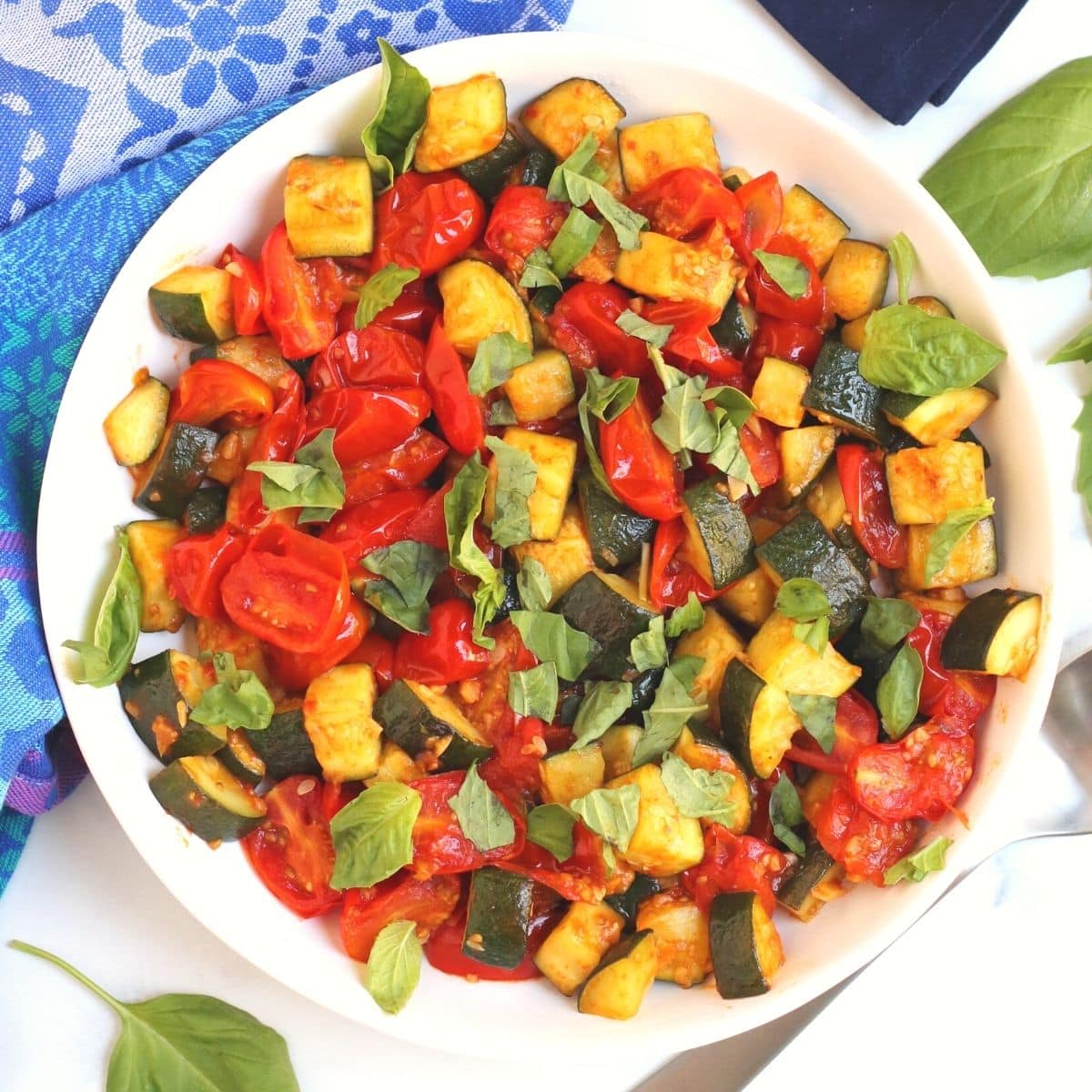 Overhead of white bowl containing sauteed zucchini and tomatoes garnished with fresh basil leaves