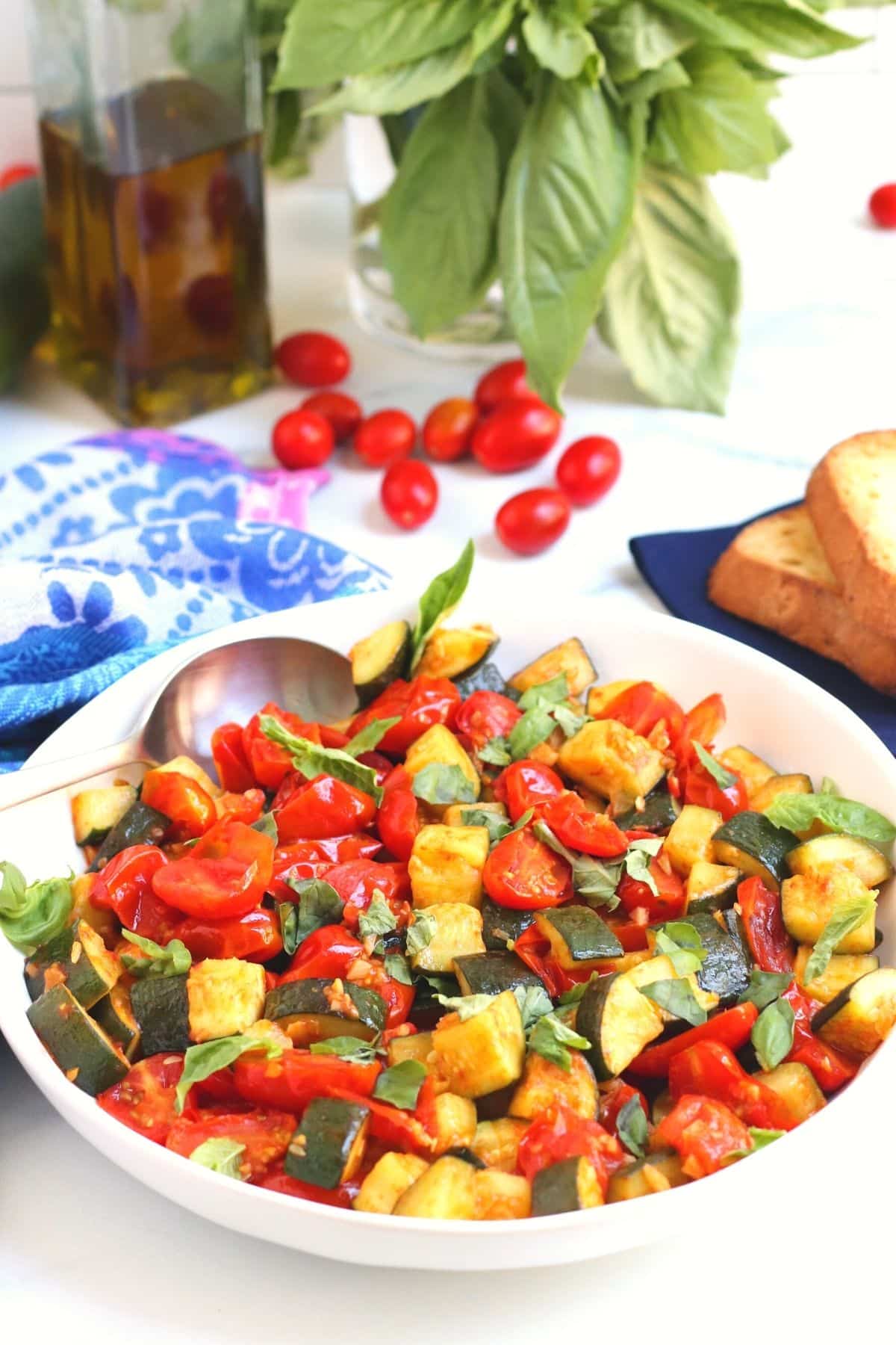 Finished side dish in a white bowls with a serving spoon in it with bottle of olive oil, grape tomatoes, and fresh basil in the background