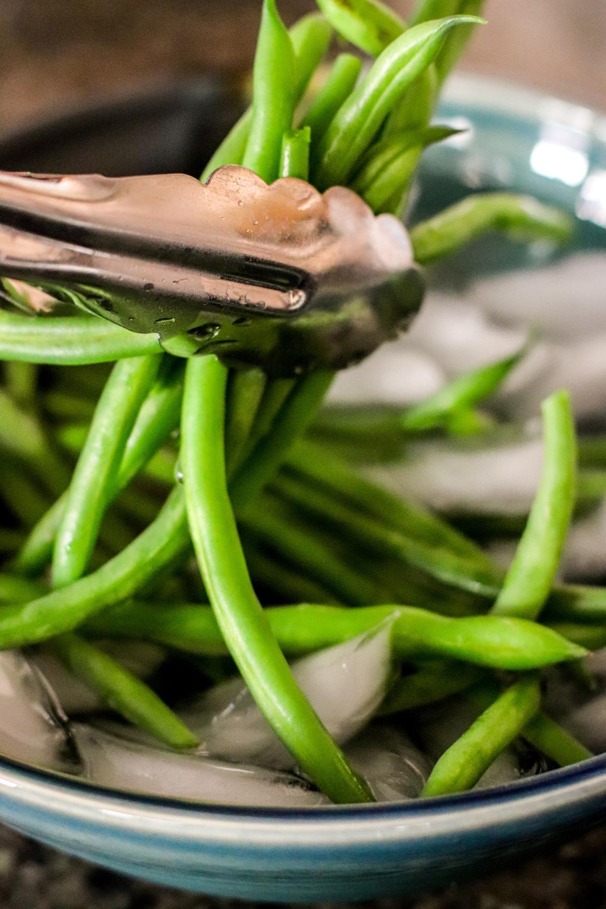 Using tongs to transfer the green beans from the boiling water to the bowl of ice water.