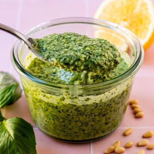 Spoon in a bowl of vegan pesto sauce surrounded by basil leaves, pine nuts, and half a lemon.