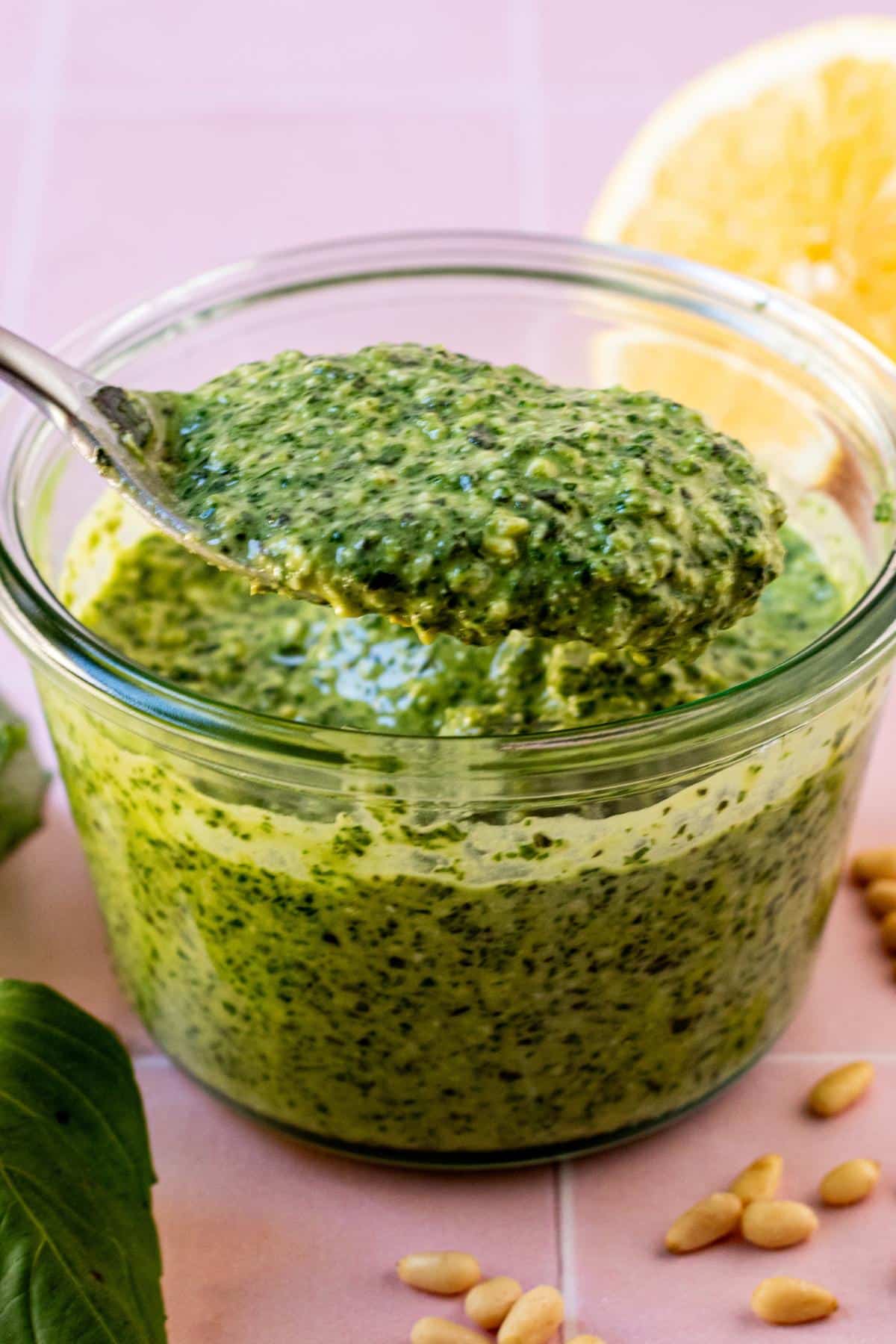 Spoon in a jar of vegan pesto sauce surrounded by fresh basil leaves, pine nuts, and a cut lemon.