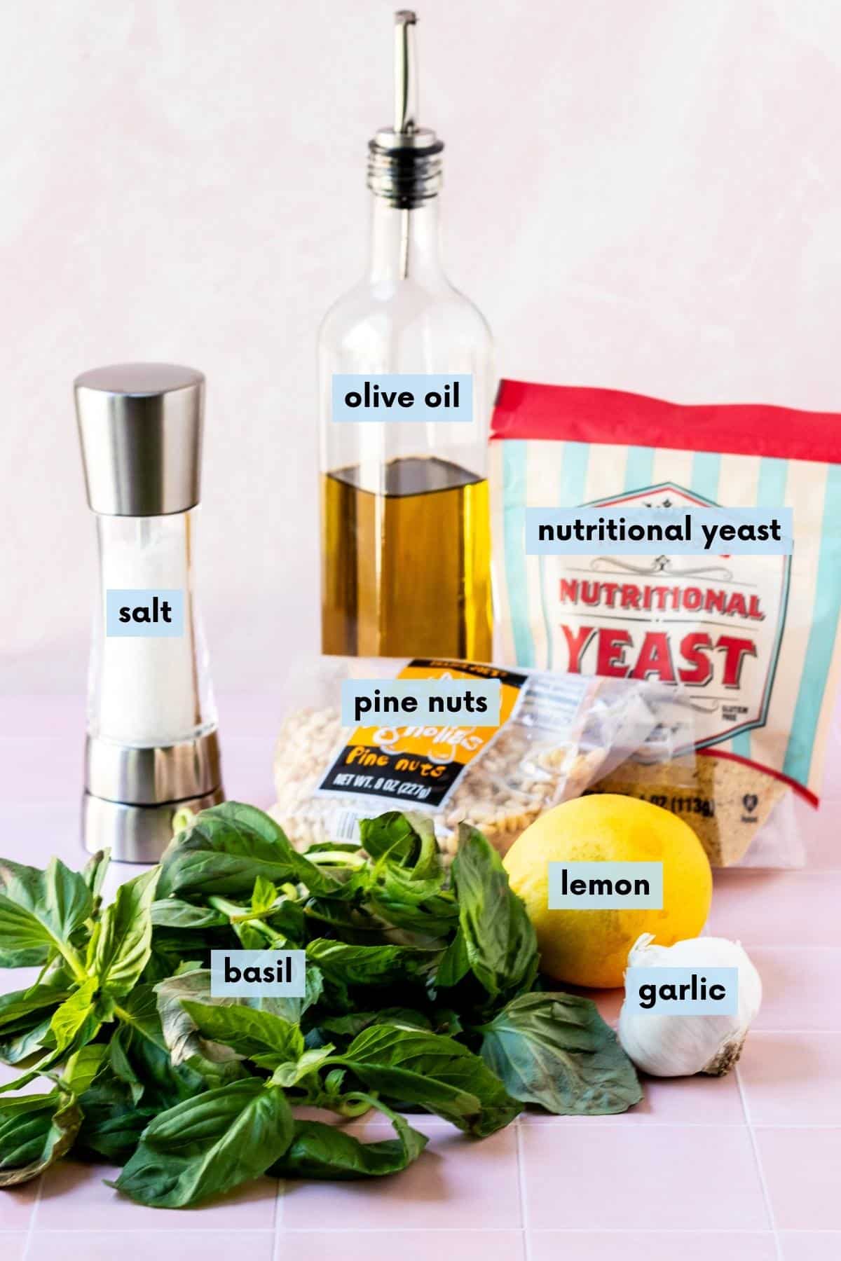 Fresh basil, a lemon, head of garlic, bag of pine nuts, salt shaker, bottle of olive oil, and bag of nutritional yeast.