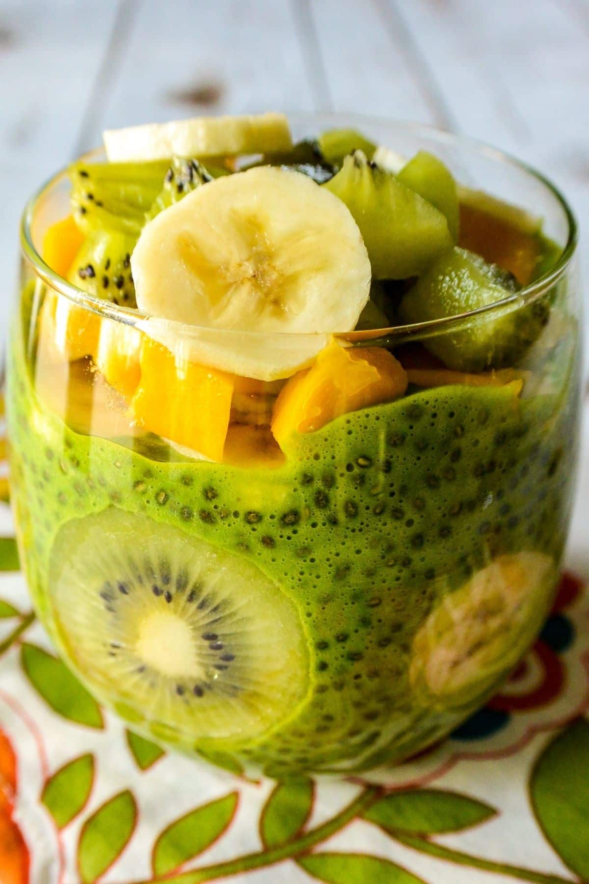 Green chia seed pudding in a clear glass layered and topped with sliced kiwi, mango, and banana