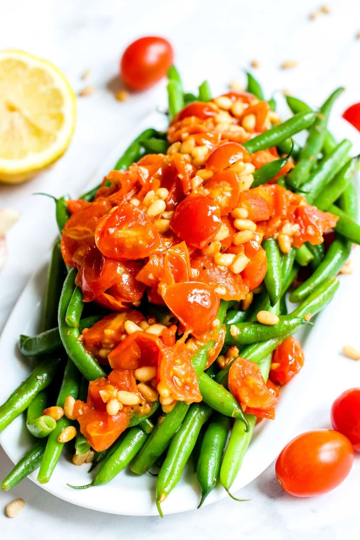 Platter of green beans topped with cooked cherry tomatoes and pine nuts with a cut lemon in the background