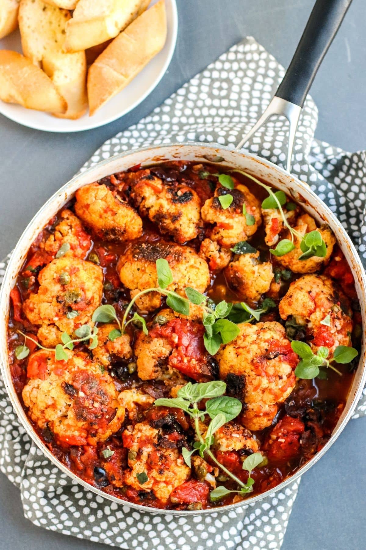 Overhead view of a skillet of charred cauliflower florets in Puttanesca sauce topped with sprigs of fresh oregano with a plate of crusty warm bread off to the side.