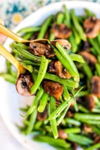 Serving spoons holding green beans and mushrooms above the serving bowl.