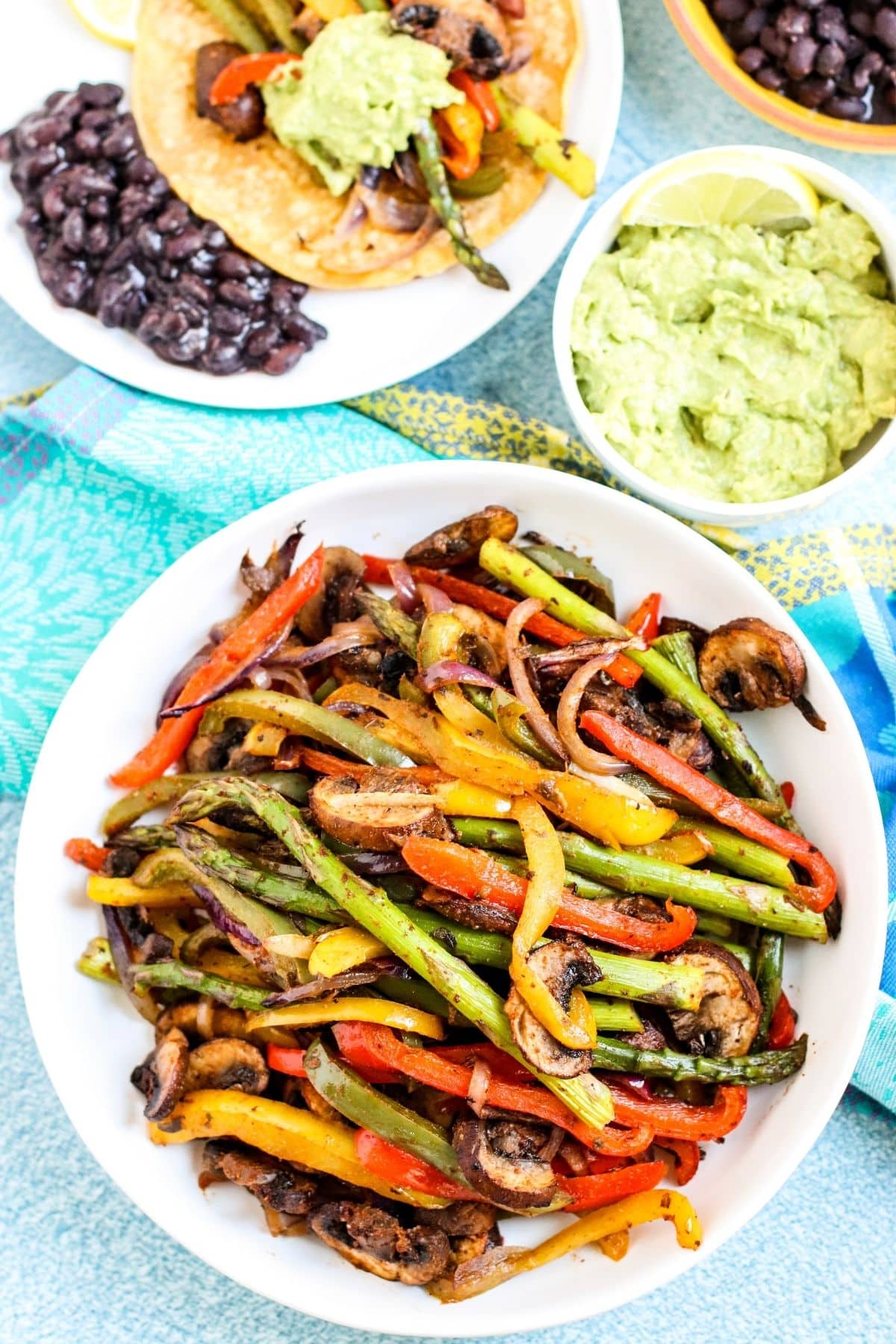 Platter of veggie fajitas, bowl of guacamole, and plate with black beans and taco