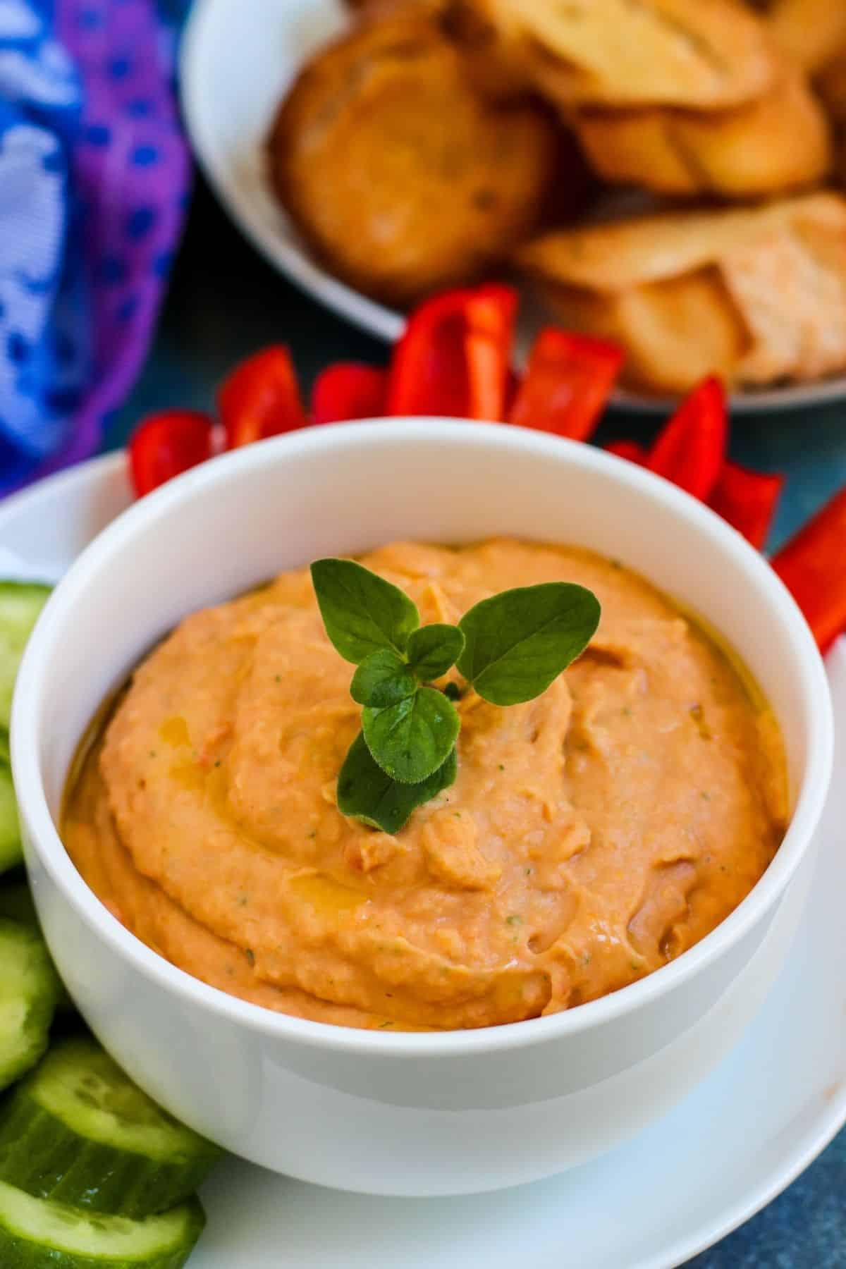 Bowl of dip garnished with a sprig of fresh oregano with raw vegetables and toasted pieces of baguette around it.