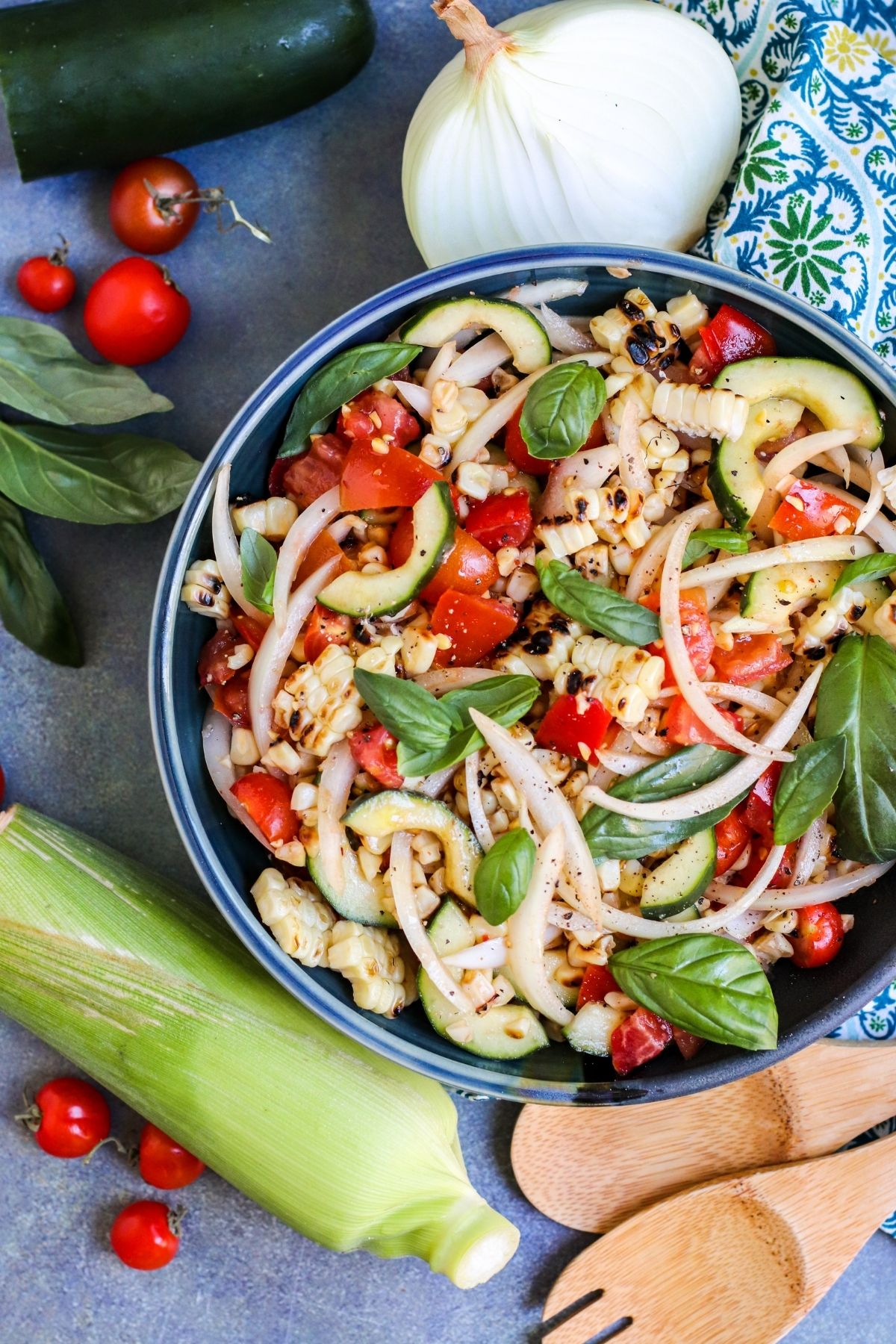 Bowl of salad surrounded by an onion, a cucumber, cherry tomatoes, fresh basil, and a cob of corn