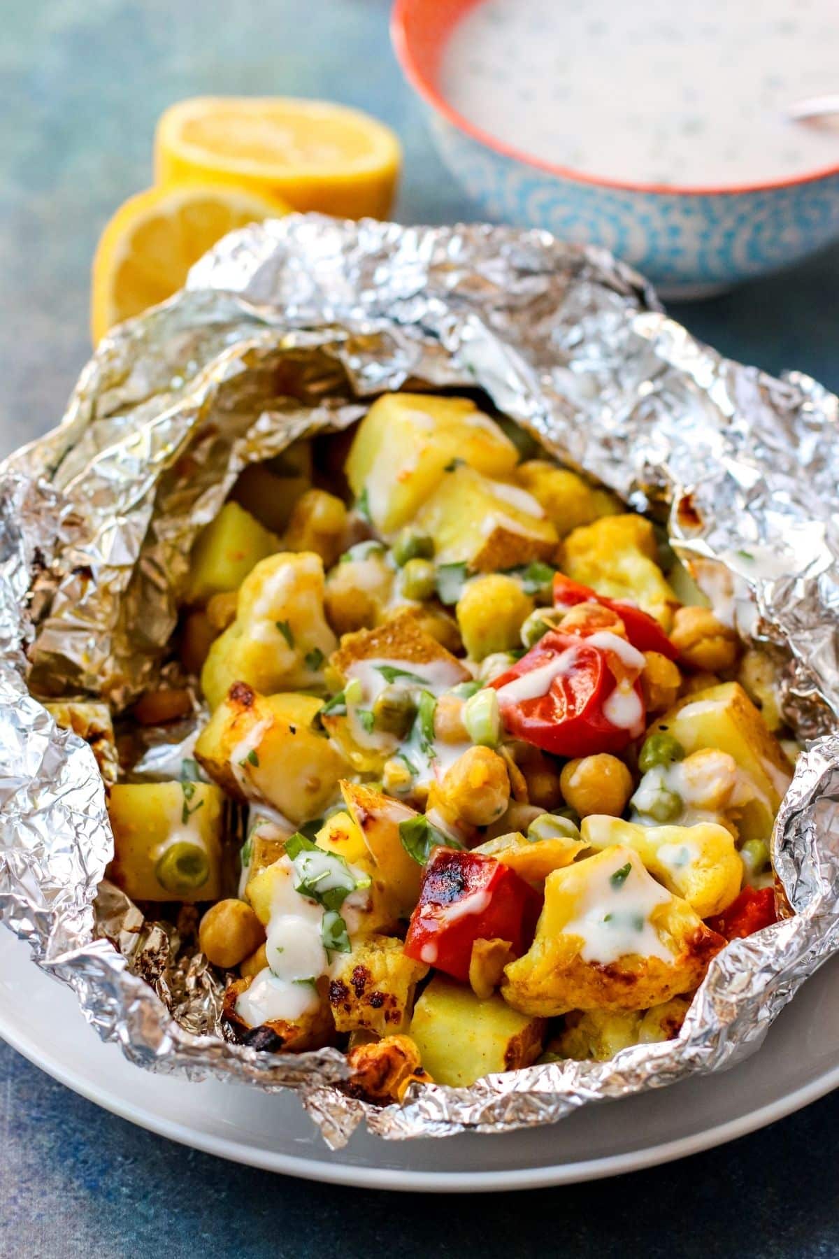 Foil packet containing potatoes, cauliflower, chickpeas, peas, and tomatoes open on a plate and drizzled with yogurt sauce