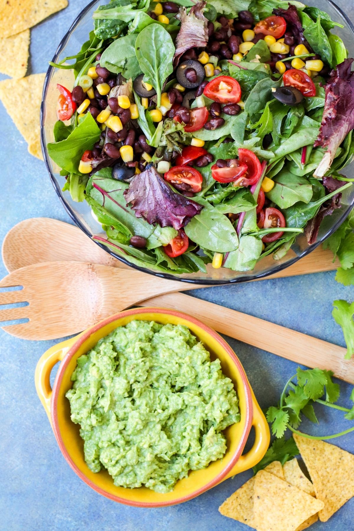 Large bowl of salad ingredients next to salad serving spoons and small bowl of guacamole