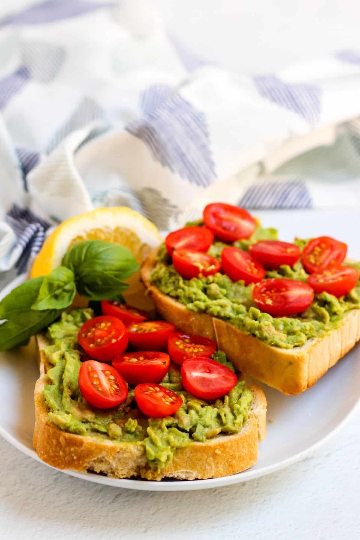 Finished slices of avocado toast on a plate topped with halved cherry tomatoes and garnished with sprig of basil and lemon wedge