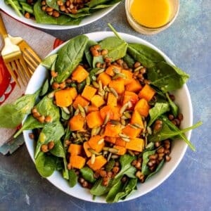 Bowl of spinach salad topped with butternut squash cubes, lentils, and pumpkin seeds.
