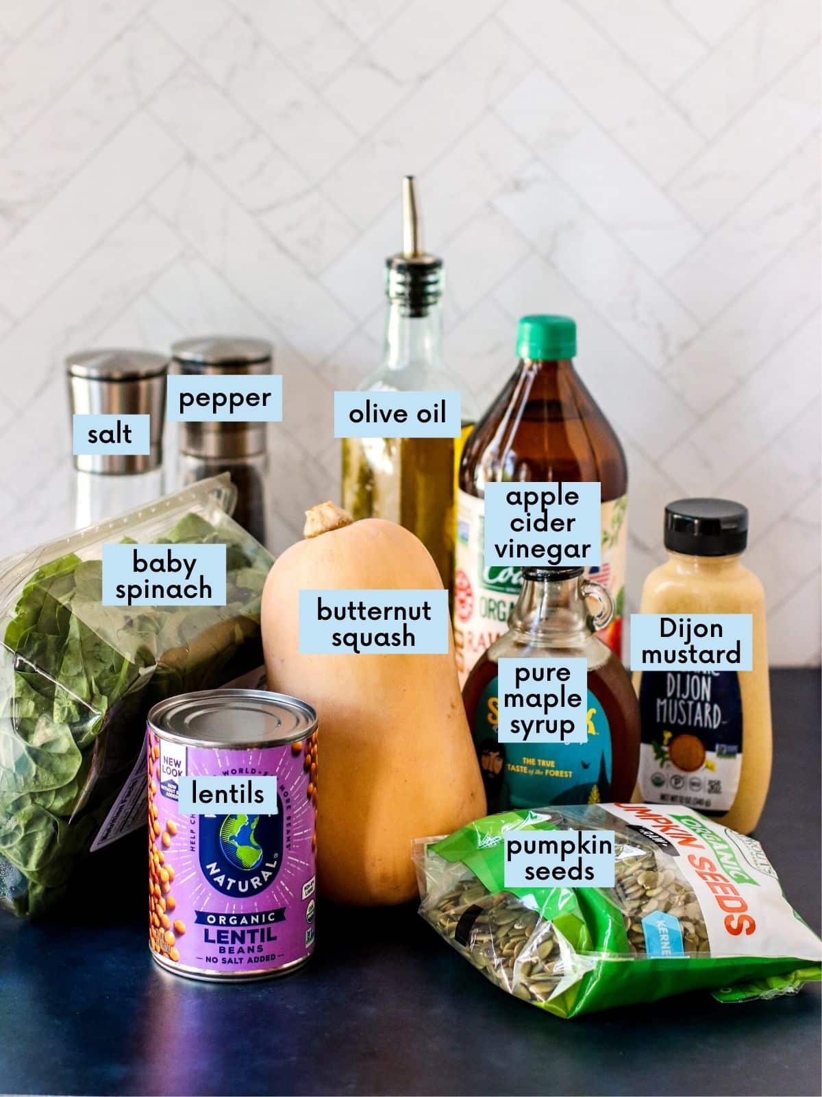 Labeled ingredients needed to make this Butternut Squash Spinach Salad: can of lentils, container of baby spinach, butternut squash, bag of pumpkin seeds, bottle of pure maple syrup, bottle of apple cider vinegar, bottle of Dijon mustard, bottle of olive oil, and salt and pepper.