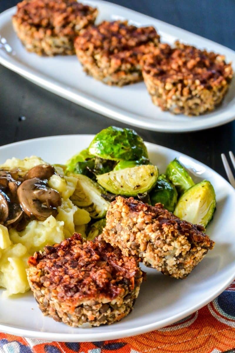 Lentil muffins on a dinner plate with mashed potatoes, mushroom gravy, and Brussels sprouts with platter of meatloaf muffins in the background.