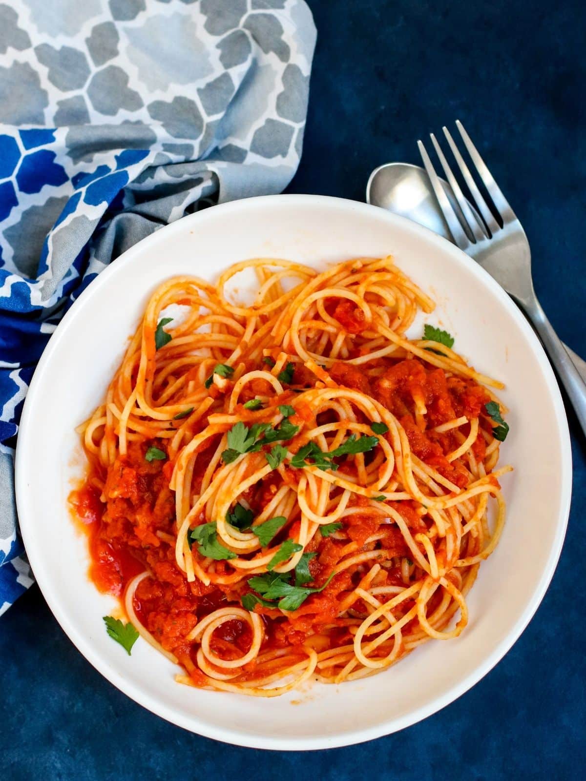 Overhead of bowl of spaghetti with pumpkin marinara sauce and fresh parsley next to a spoon, fork, and patterned blue and gray napkin.