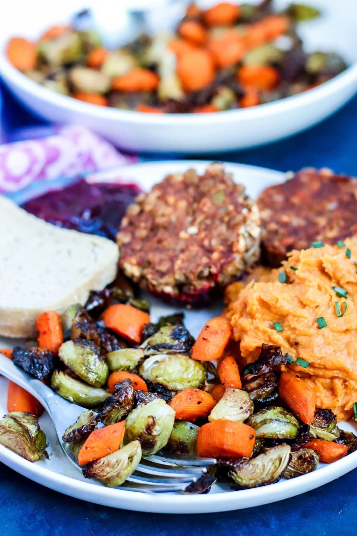 Roasted Brussels sprouts and carrots on a dinner plate with mashed sweet potatoes, lentil meatloaf muffins, cranberry sauce, and bread with bowl of vegetables in the background.
