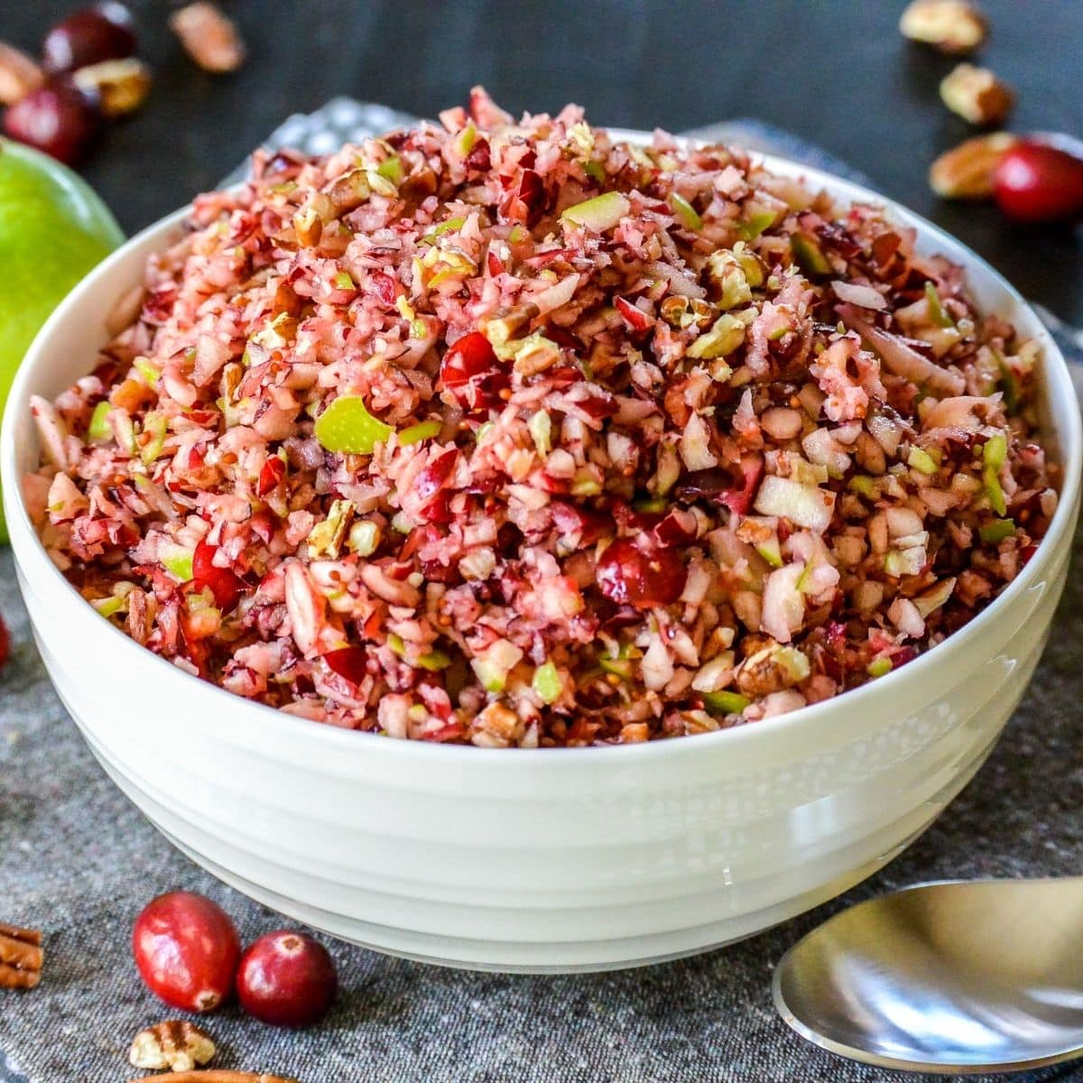 Bowl of relish surrounded by raw cranberries, a green apple, and pecans.