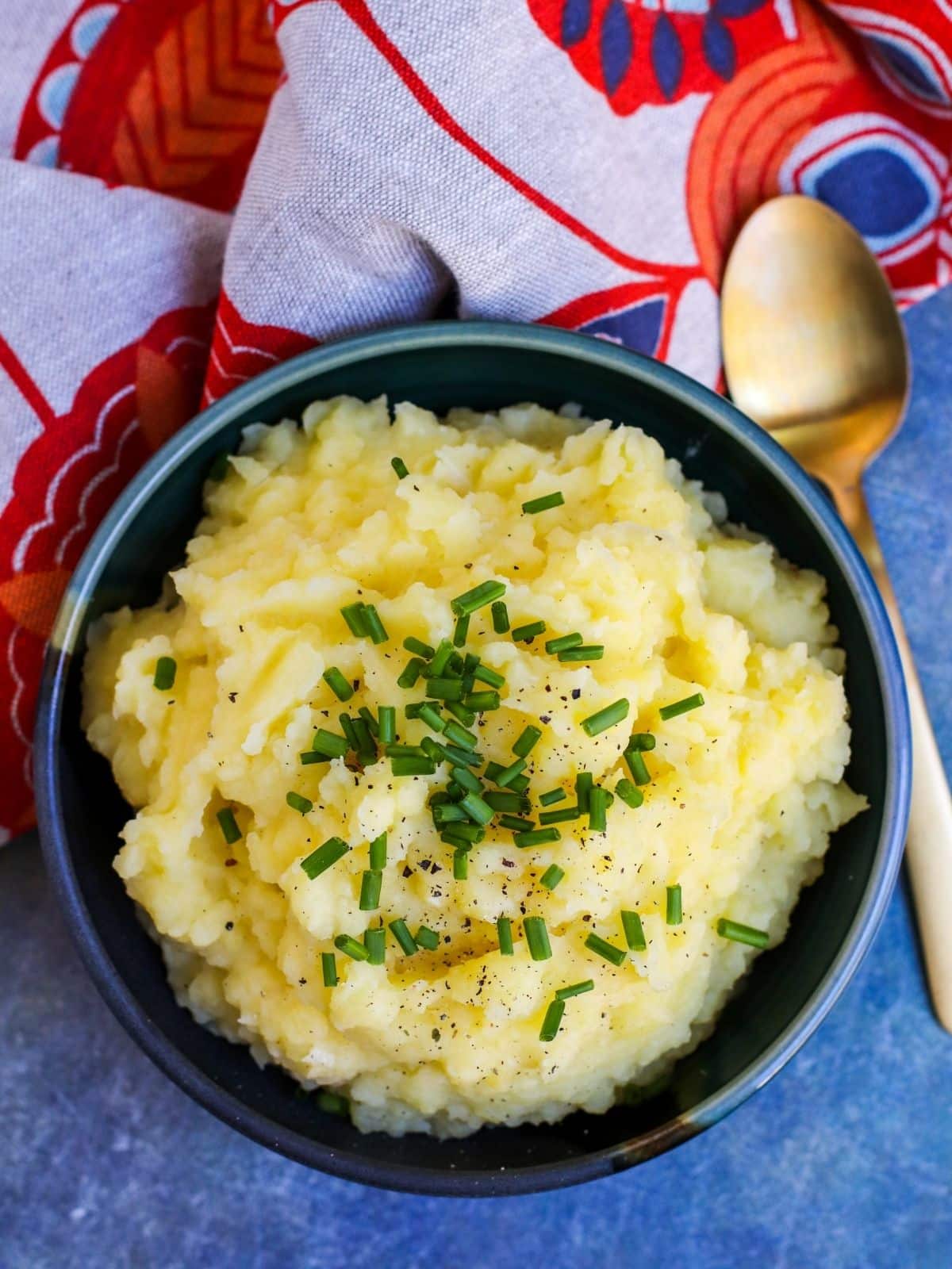Bowl of mashed potatoes topped with chives with a gold spoon and orange and gray napkin.