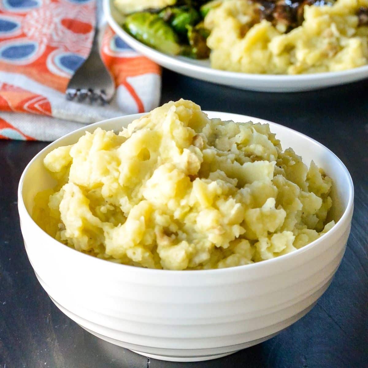 Bowl of mashed potatoes with dinner plate in the background.