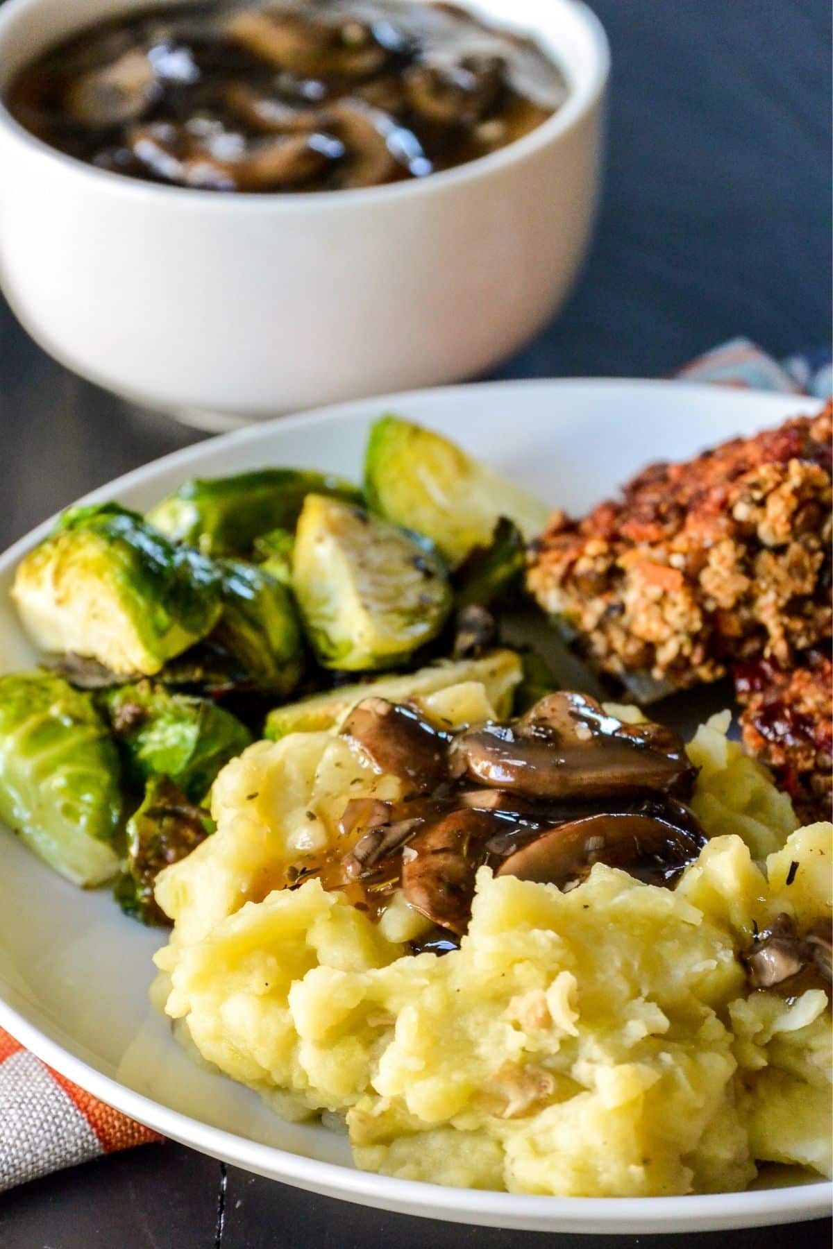 Dinner plate with mashed potatoes, mushroom gravy, roasted Brussels sprouts, and lentil meatloaf with bowl of gravy in the background.