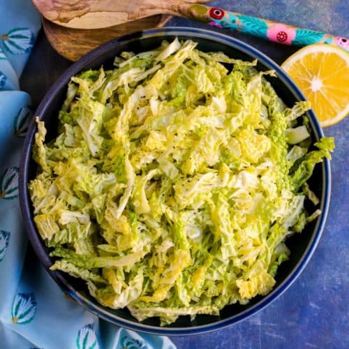 Bowl of cabbage salad with cut lemon and serving utensils.