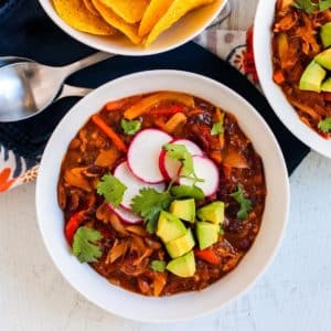 Bowl of chili topped with radish slices, fresh cilantro, and avocado cubes next to spoons and a bowl of tortilla chips.