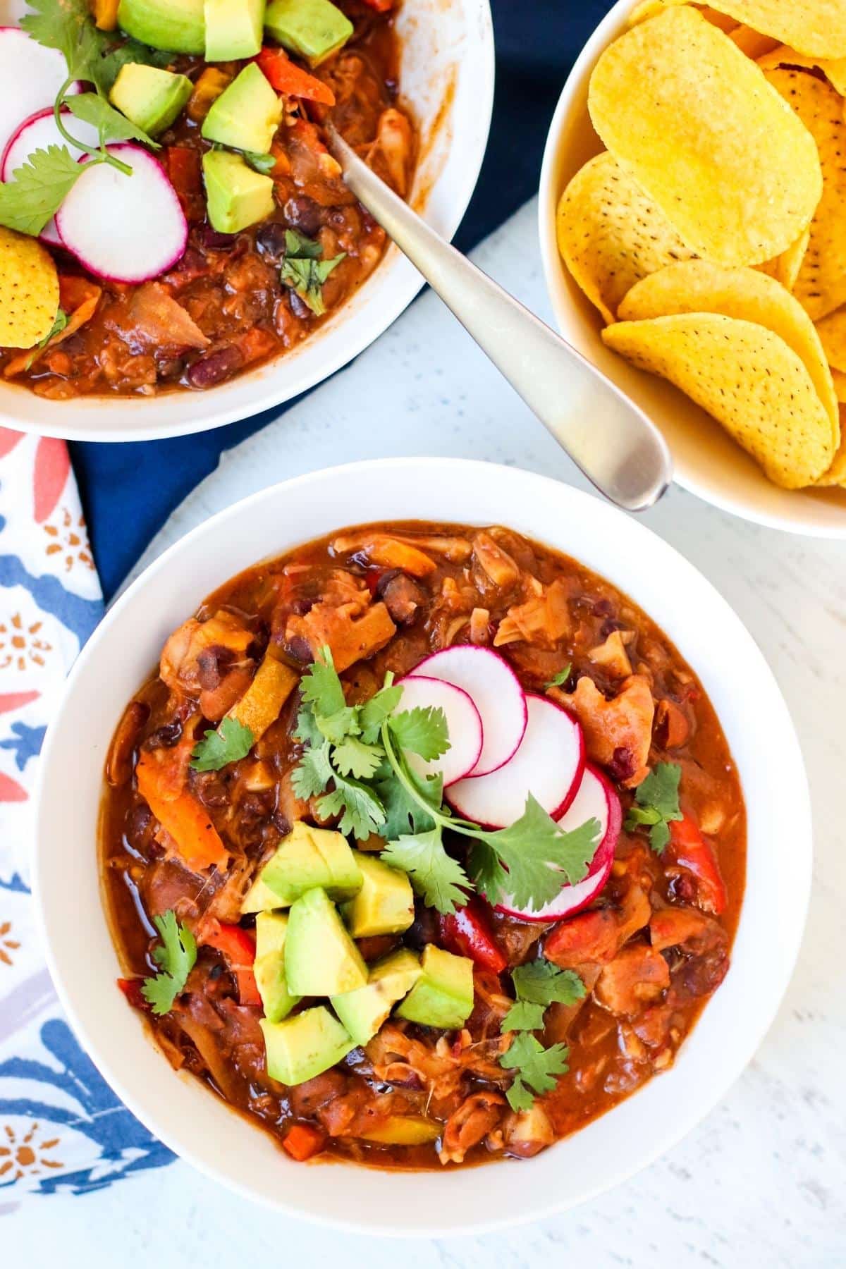 Bowls of jackfruit chili topped with avocado, cilantro, and radish slices and bowl or tortilla chips.