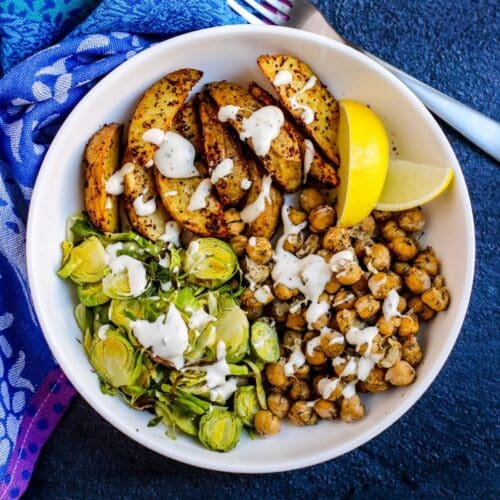 Roasted potato wedges, chickpeas, and Brussels sprouts drizzled with sauce and garnished with lemon wedges.