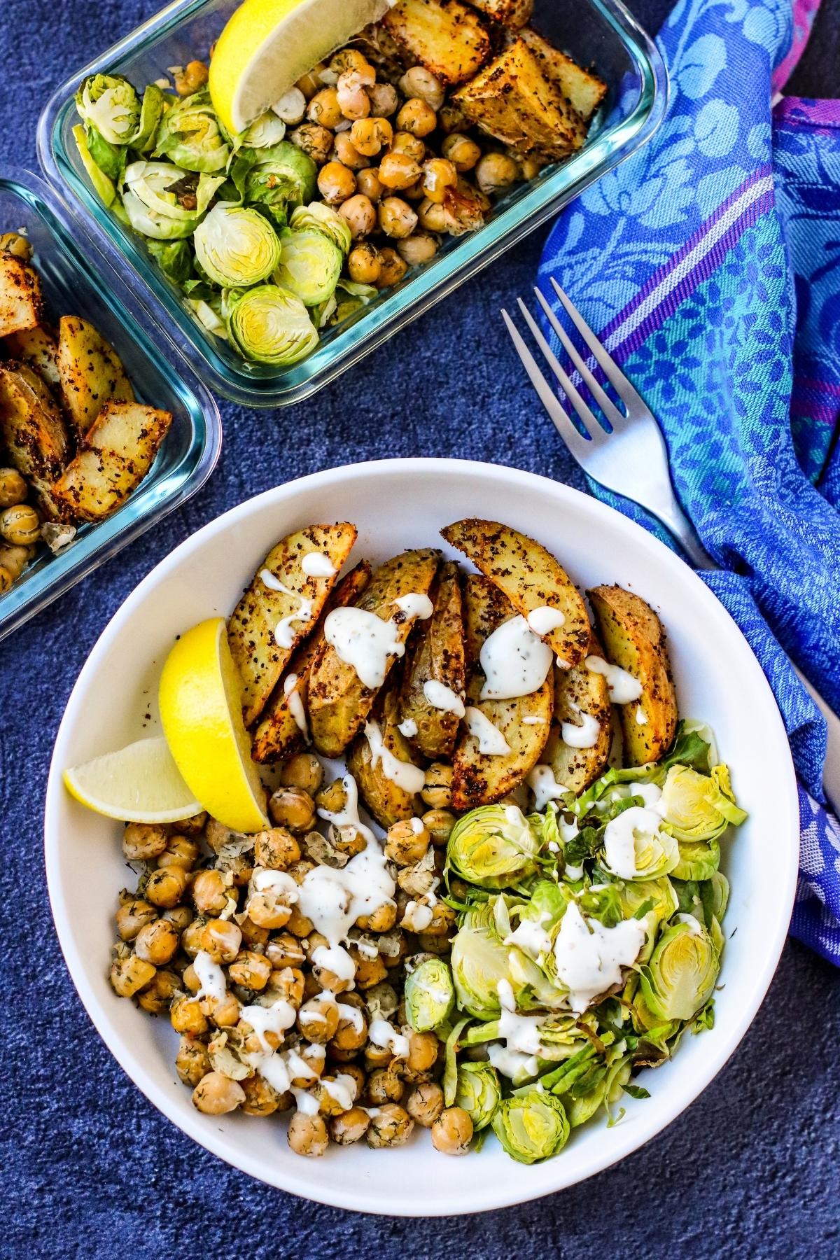 Chickpea Bowl with potatoes and Brussels sprouts drizzled with sauce with meal prep containers in the background.