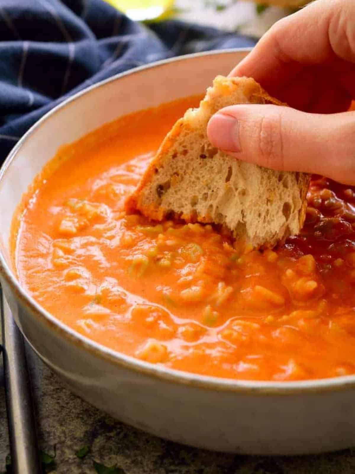 Hand dipping crusty bread into a bowl of tomato soup.