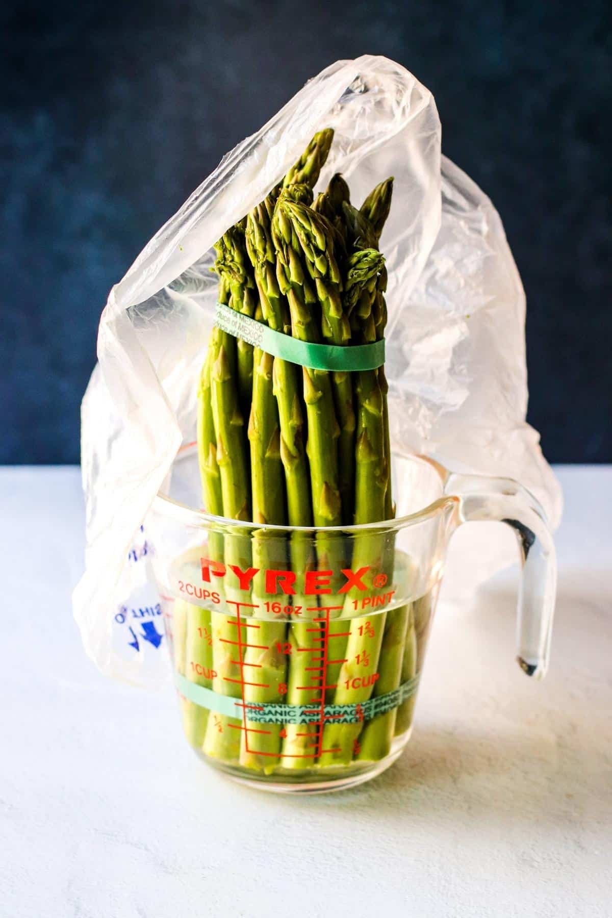 Bunch of asparagus in a Pyrex cup with water covered with a plastic bag.