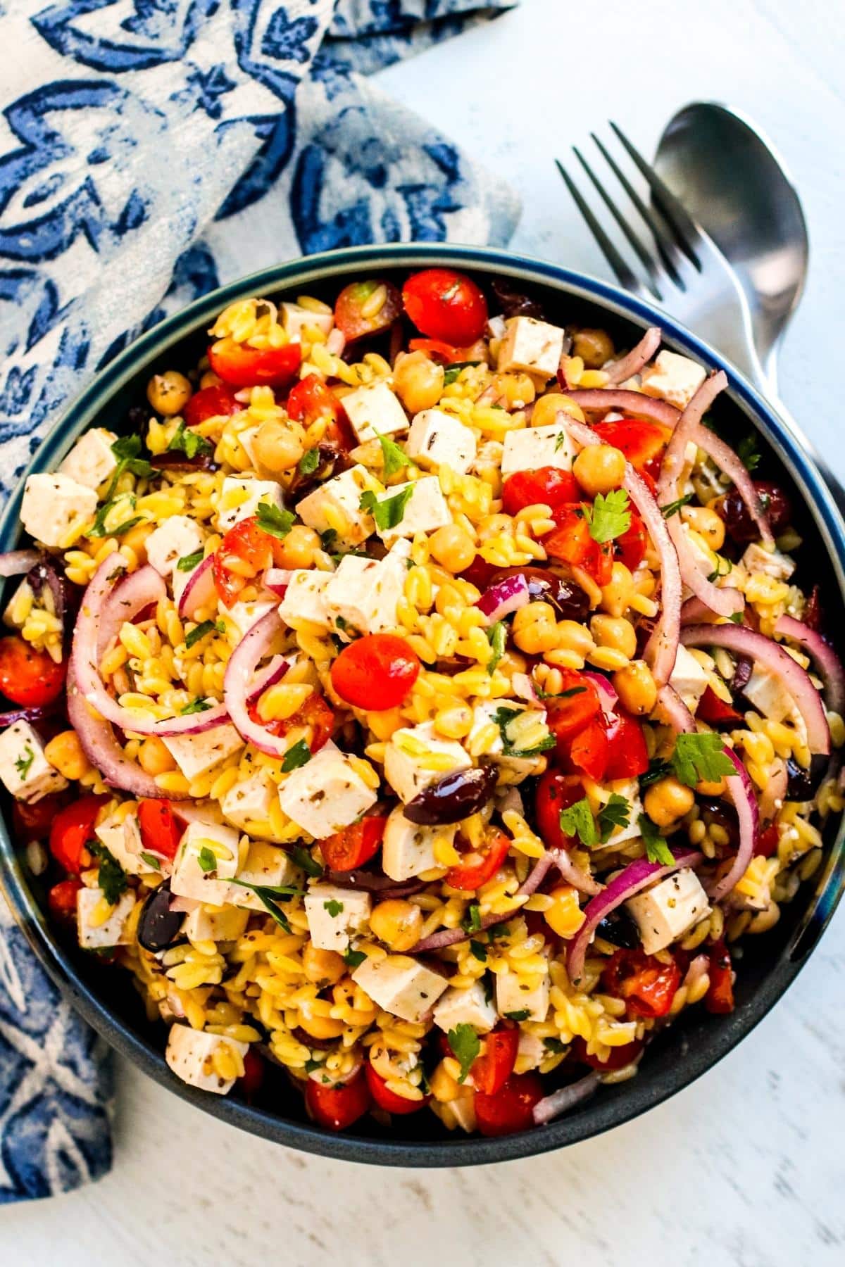 Bowl of Mediterranean Vegan Orzo Salad next to serving utensils and blue and white napkin.