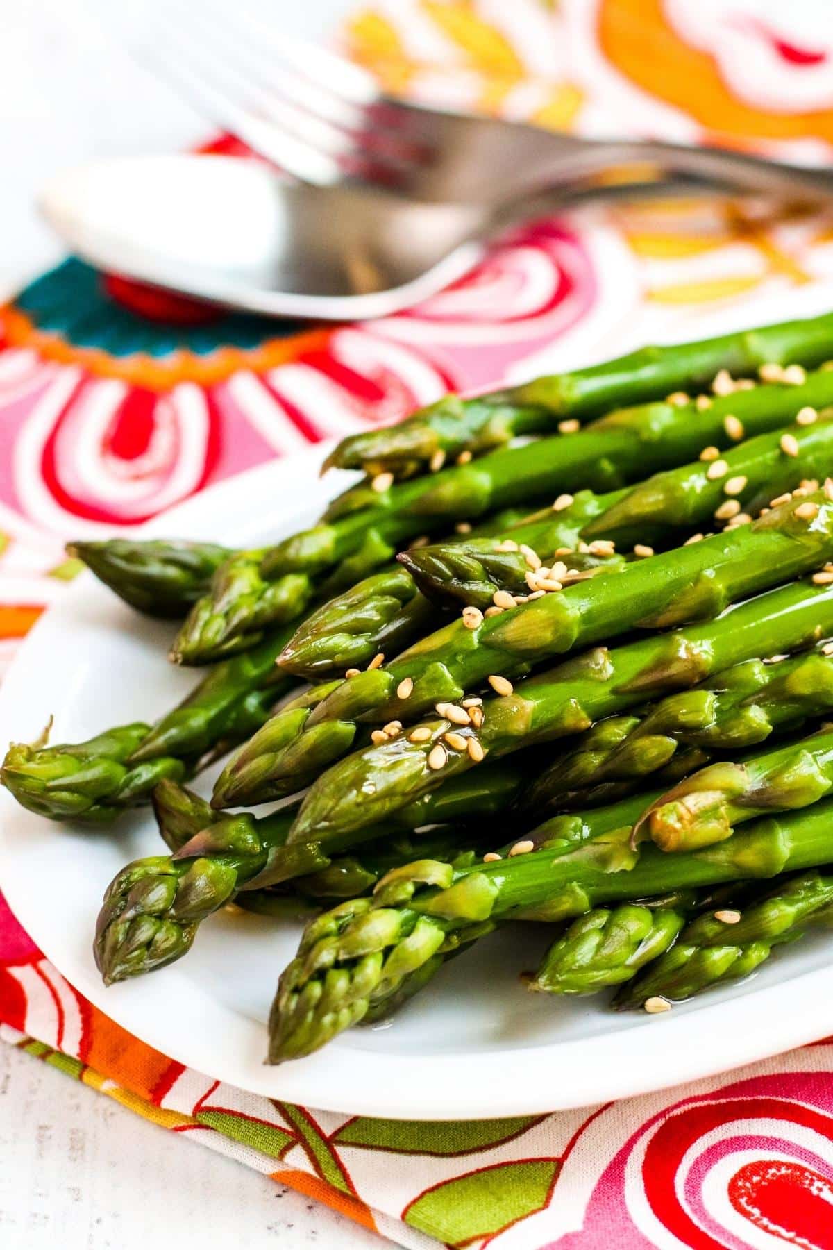 Platter of sesame seed topped marinated asparagus and serving utensils on a floral napkin.