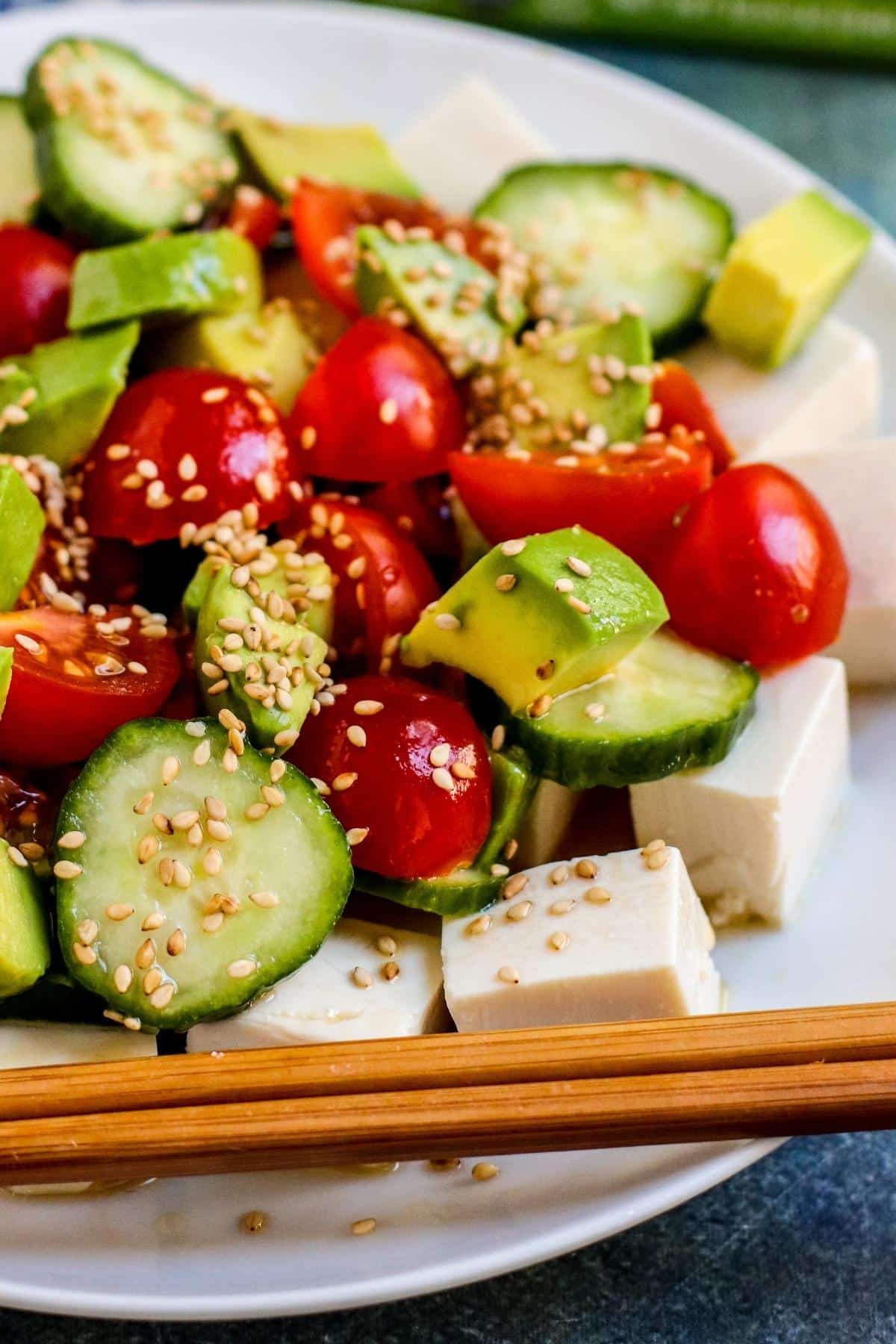 Close up of sliced cucumbers, quartered cherry tomatoes, chunks of avocado, and cubes of tofu topped with toasted sesame seeds next to wooden chopsticks.