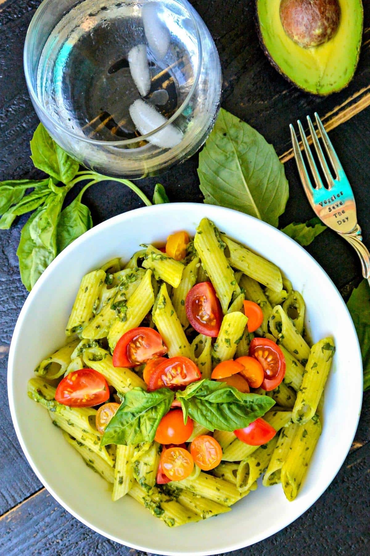 Overhead image of bowl of pasta, water glass, and fork with basil leaves and half an avocado.