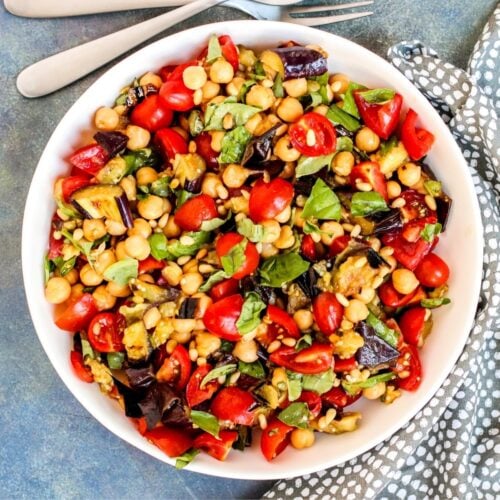 Serving bowl of eggplant salad with serving utensils and gray napkin.