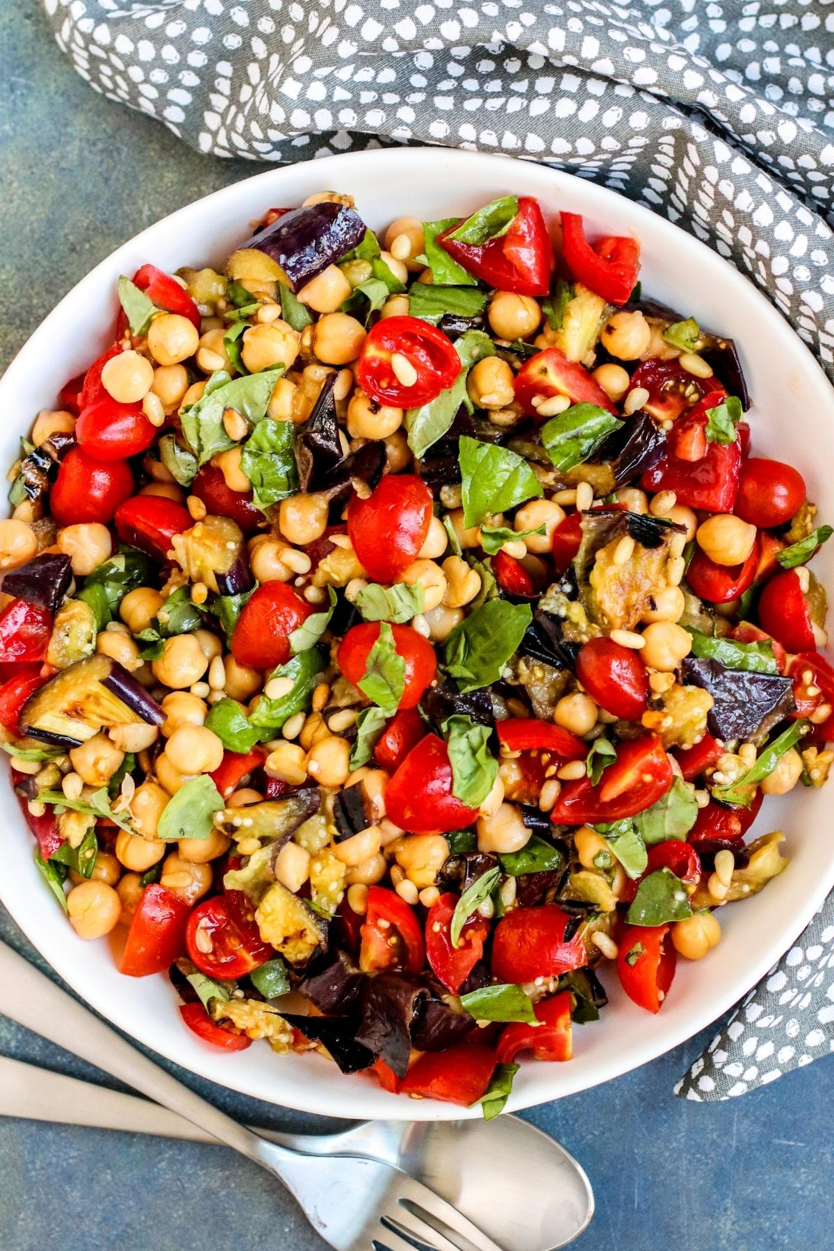 Serving bowl of Mediterranean Grilled Eggplant Salad with serving utensils and gray napkin.